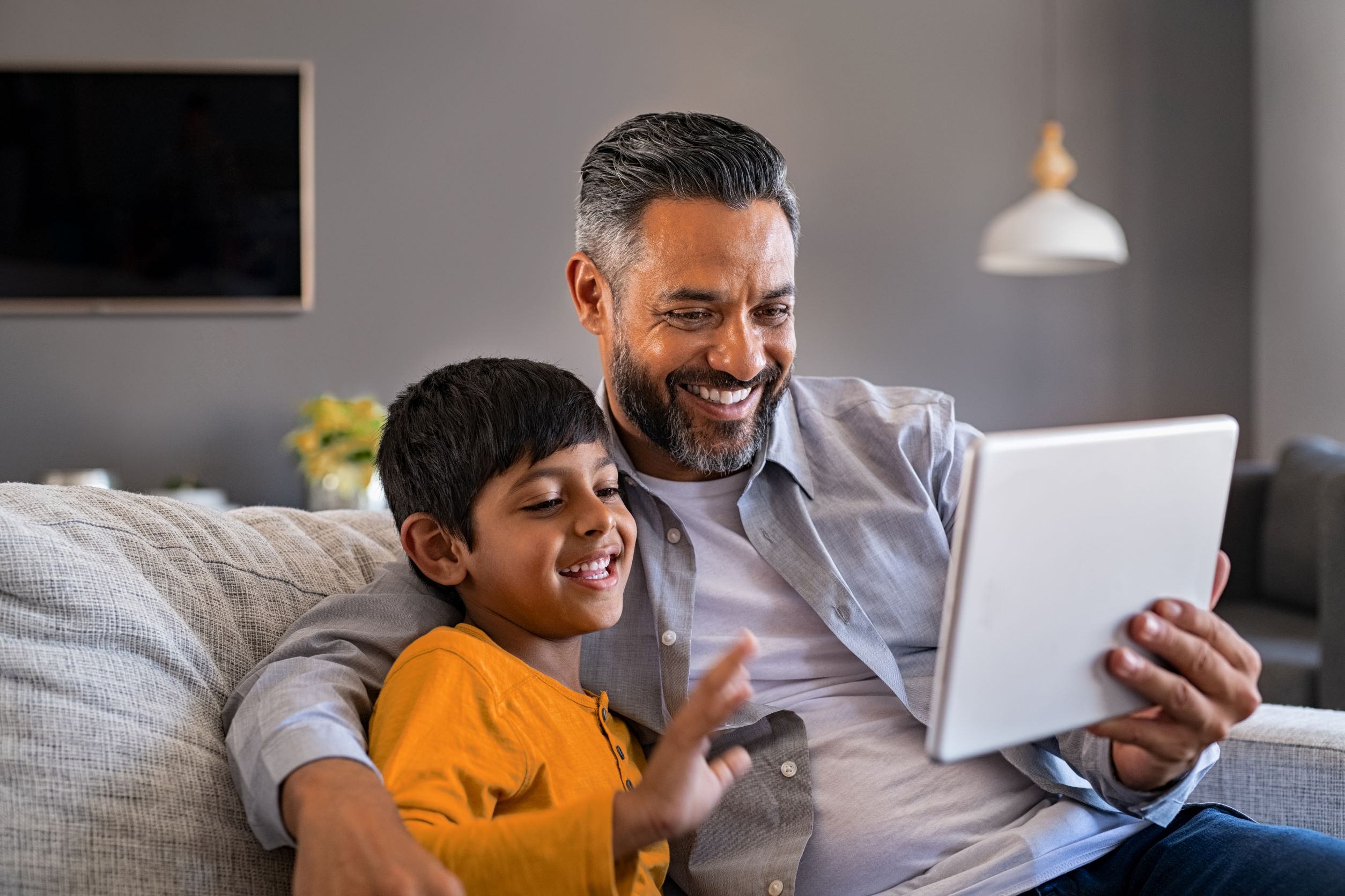 A father and son seating on their couch, the dad is holding up a tablet and they are both looking at the tablet screen